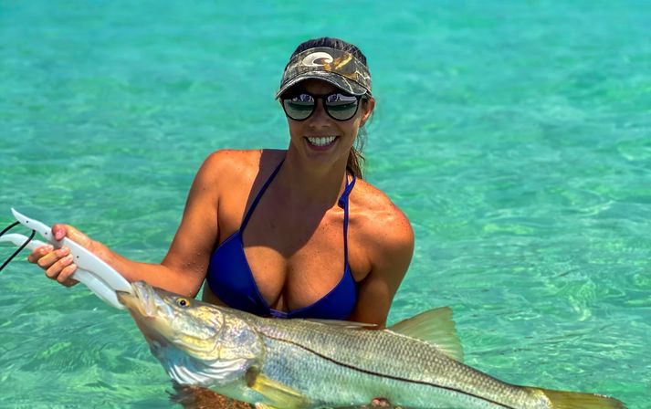 Smiling angler in a blue bikini and visor holding a large snook over clear turquoise tropical flats during saltwater fishing