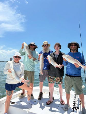Five anglers on a small boat holding redfish after a successful saltwater fishing trip on calm coastal waters under a bright blue sky.