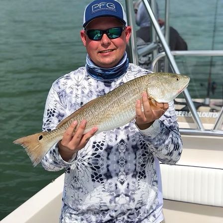 Angler on a boat holding a healthy red drum (redfish) with a black tail spot over calm coastal waters.