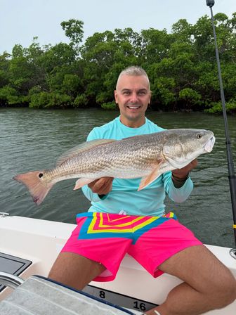 Smiling angler on a boat holding a large redfish (red drum) over coastal mangrove-lined water, wearing a turquoise shirt and bright neon-pink geometric swim trunks.