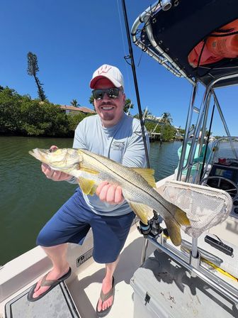 Angler in sunglasses and a baseball cap proudly holding a large snook on a sunny boat ride through a mangrove-lined Florida canal