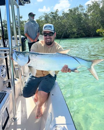 Grinning angler on a boat holding a large silver tarpon over clear turquoise flats by a mangrove shoreline — sunny saltwater fishing catch in shallow coastal waters.