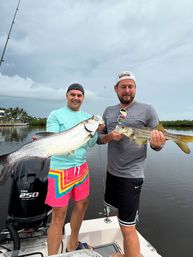 Two anglers on a small boat holding a large tarpon and a snook over a calm mangrove-lined coastal inlet under a cloudy sky, celebrating a big catch.
