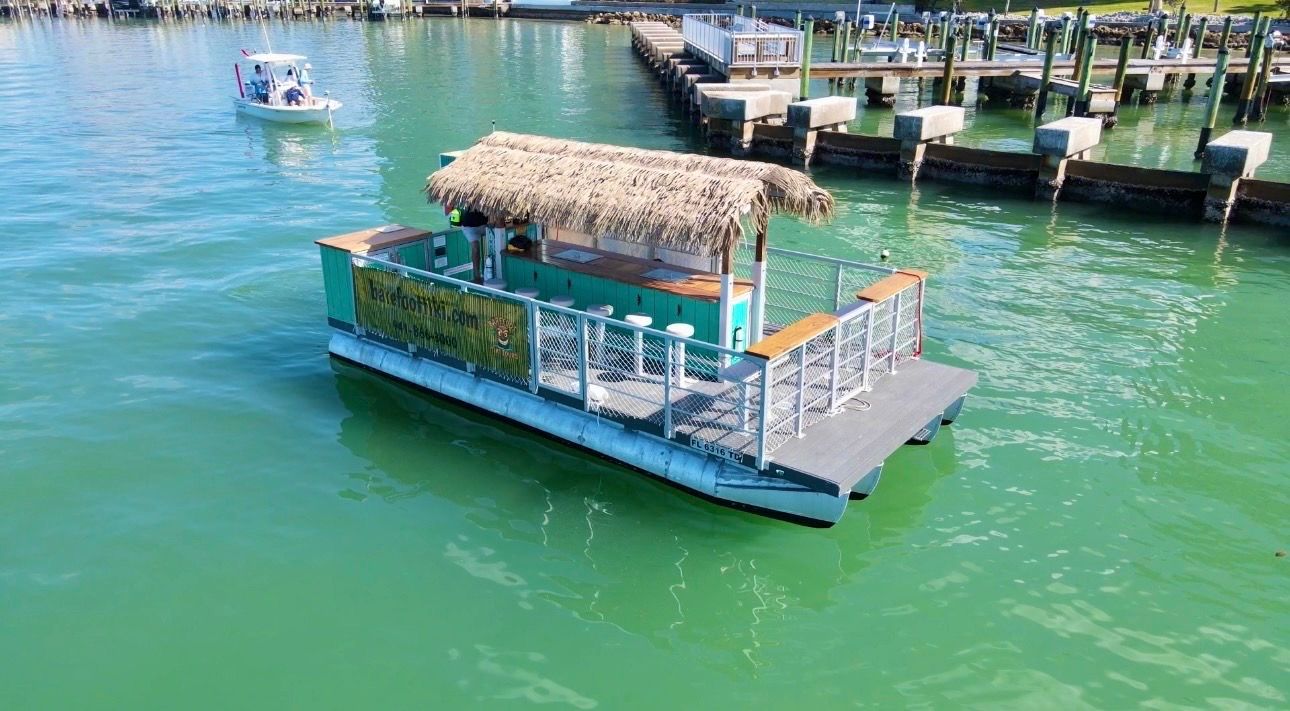 Floating tiki-bar pontoon with a straw thatched roof and bar stools drifting near a marina pier in clear turquoise coastal waters, a small motorboat nearby.