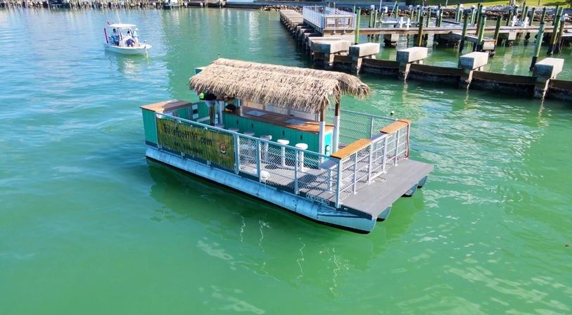Floating tiki-bar pontoon with a straw thatched roof and bar stools drifting near a marina pier in clear turquoise coastal waters, a small motorboat nearby.