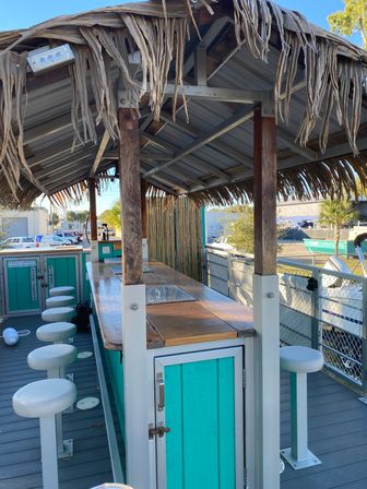 Sunny outdoor tiki-style bar with turquoise wooden counter, white swivel bar stools and thatched roof on a marina dock, palm trees and boats visible in the background