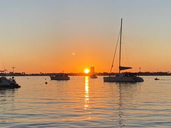 Golden sunset over a calm harbor with silhouetted sailboats and a catamaran, sunlight forming a shimmering path across the water.
