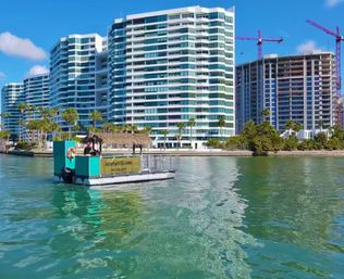 Tropical waterfront scene with a turquoise tiki-style pontoon boat floating in clear green water in front of modern white glass high-rise condos, palm trees, and construction cranes under a bright blue sky.
