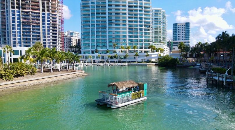Tiki-style floating bar boat cruising a green urban canal past palm-lined marina and high-rise waterfront condos under a sunny blue sky