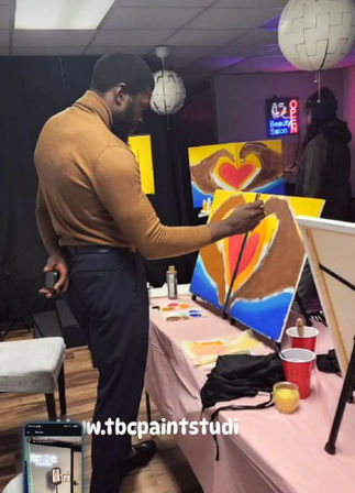 Adult painting a vibrant heart design on an easel during an indoor paint workshop, studio table with palettes, red cups and round pendant lights.