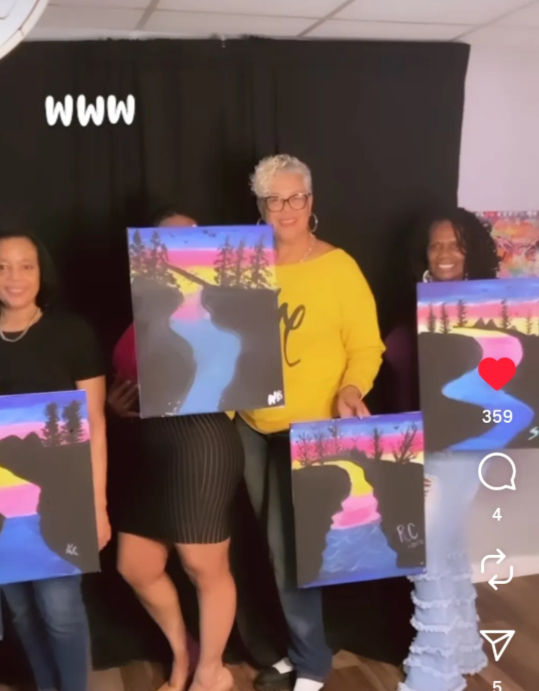 Four women in an indoor art studio proudly holding colorful sunset river landscape canvases from a group painting class — pink, yellow and blue sky above silhouetted trees and a winding river.