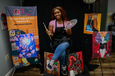 Smiling woman artist in a paint studio holding a palette and brush, seated among vibrant canvases and a banner promoting virtual, public and private paint parties.