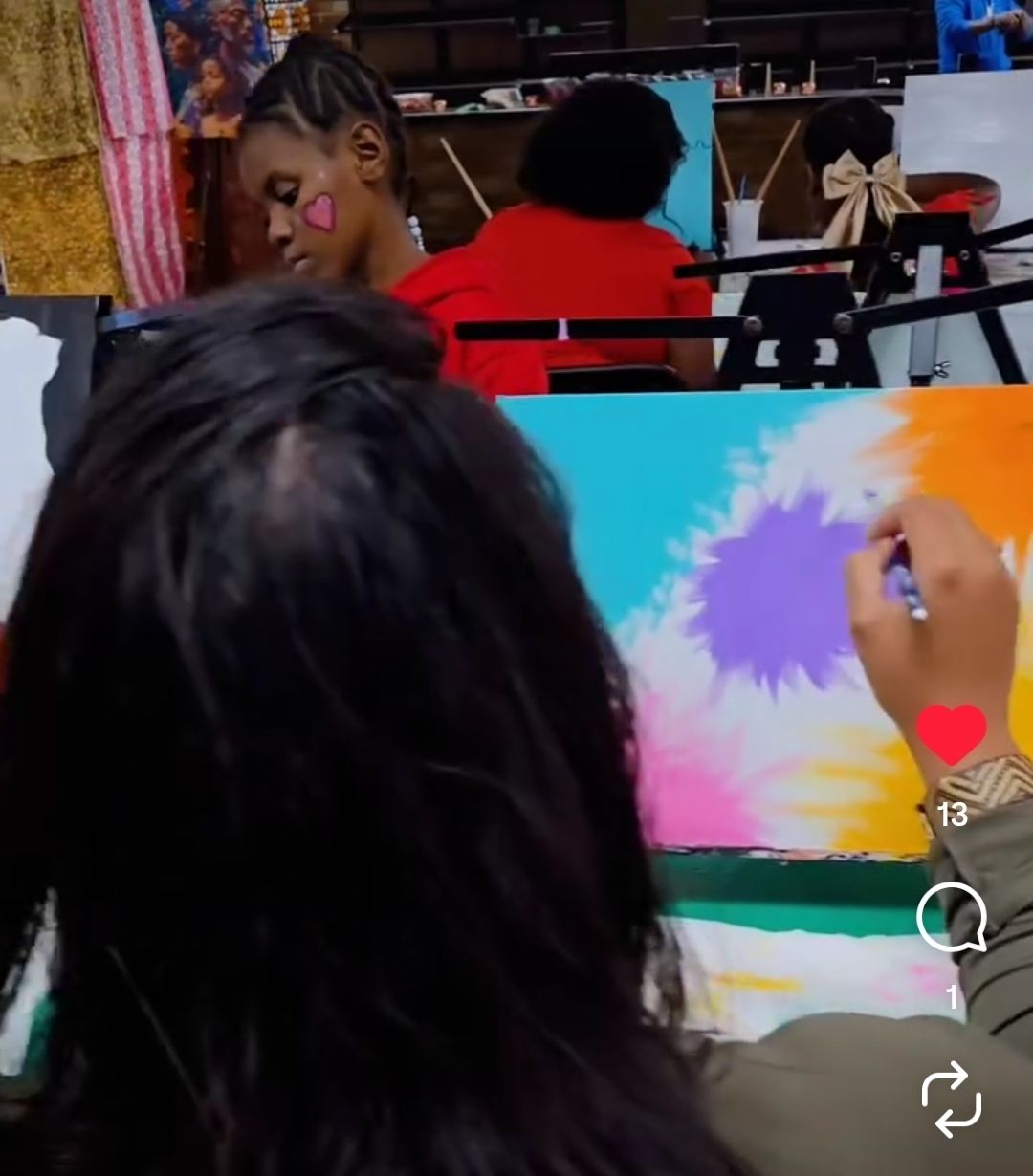 Children's painting workshop in an art studio — close-up of a person painting a bright turquoise, purple and orange canvas while another child with a pink heart face paint works at an easel.