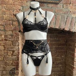 Mannequin styled in an edgy black satin and lace lingerie set with harness, garter belt and gold hardware, displayed against an exposed brick wall.