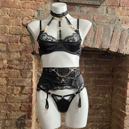 Mannequin styled in an edgy black satin and lace lingerie set with harness, garter belt and gold hardware, displayed against an exposed brick wall.