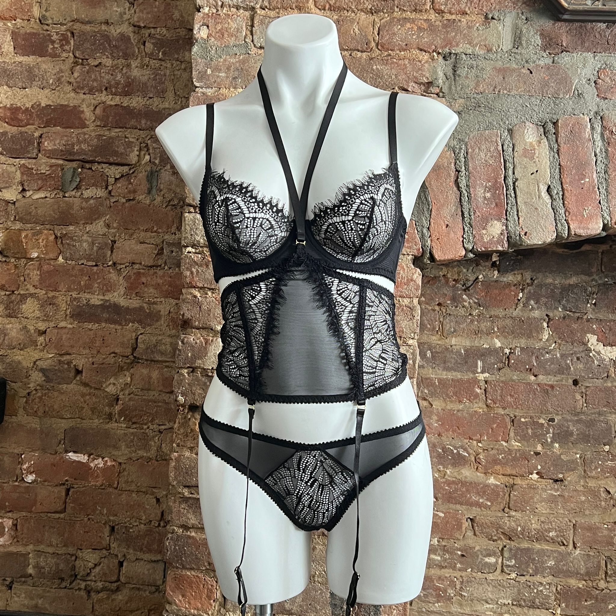 White mannequin wearing a black lace lingerie set with sheer panels and garter straps displayed against a rustic exposed brick wall