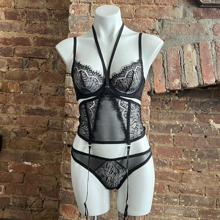 White mannequin wearing a black lace lingerie set with sheer panels and garter straps displayed against a rustic exposed brick wall