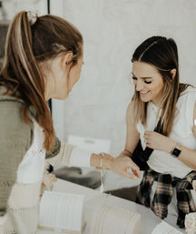 Two women smiling while jewelry shopping at a bright boutique-style table display; one woman tries on a bracelet as delicate necklaces and cuffs are arranged nearby.