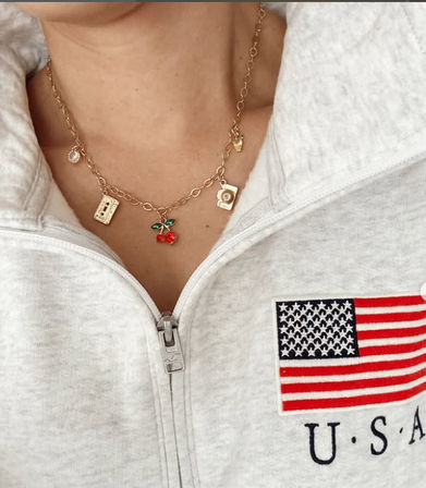 Close-up of a gold charm necklace with a red cherry pendant and assorted charms worn over a light gray zip-up sweatshirt embroidered with a U.S.A. flag.