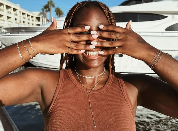 Smiling woman with braided hair and white manicure covering her eyes with stacked rings and bracelets in front of yachts at a sunny marina, wearing layered gold necklaces and a rust-colored tank top.