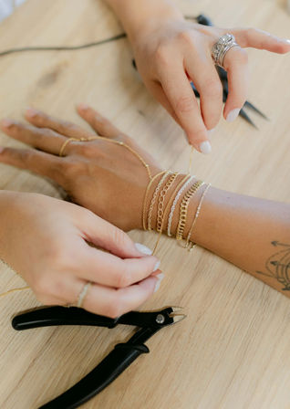 Close-up of artisan hands adjusting layered gold chain bracelets on a wrist over a wooden workbench with jewelry pliers.