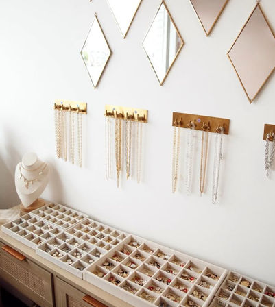 Stylish jewelry display on a white vanity — compartmentalized ring trays, a necklace bust with layered pendants, and gold wall-mounted hooks holding gold and silver chains beneath diamond-shaped mirrors.