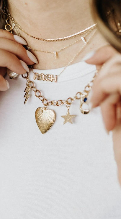 Close-up of a person’s neck wearing layered gold necklaces over a white tee — delicate chains plus a chunky link charm necklace with a heart, star and lightning charms and a letter-name charm, with stacked rings visible.