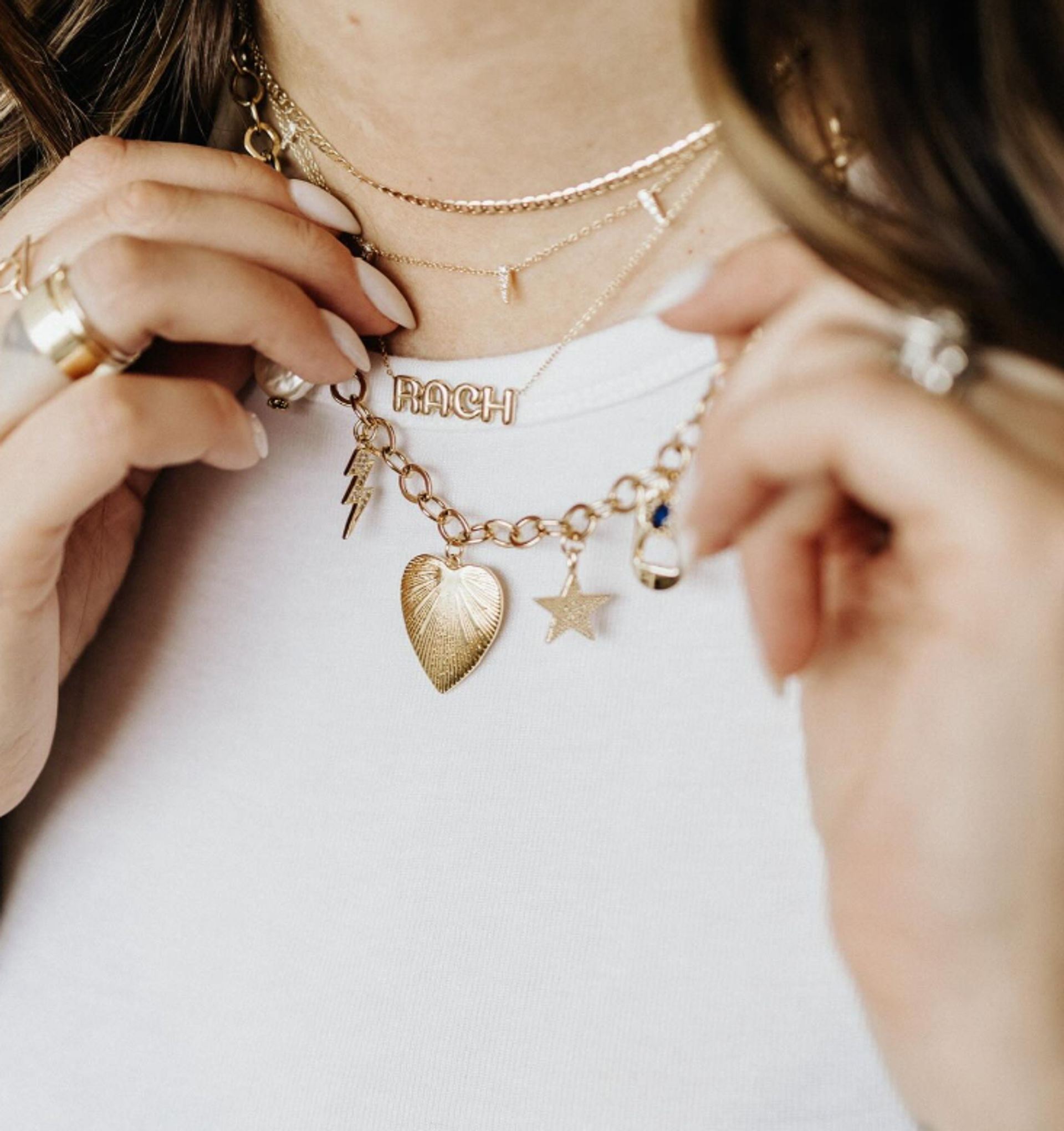 Close-up of a person’s neck wearing layered gold necklaces over a white tee — delicate chains plus a chunky link charm necklace with a heart, star and lightning charms and a letter-name charm, with stacked rings visible.