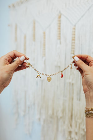 Close-up of hands holding a gold charm bracelet with a heart, red football and small pendants against a boho macrame wall — stylish jewelry product shot.