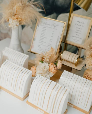 Boho-chic bridal jewelry display with delicate gold chain bracelets and necklaces on white curved stands, framed menu cards, and dried pampas and peach florals on a neutral tabletop.