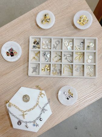 Top-down view of assorted gold and silver jewelry charms, pendants and charm bracelets arranged in a divided display tray and round stone dishes on a light wood tabletop