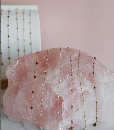Delicate gold-chain beaded bracelets with tiny multicolored gemstone beads draped over a large rose quartz slab on a soft pink backdrop.