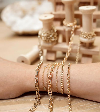 Sparkling layered gold chain bracelets on a wrist with extra chains wrapped on wooden spools in a warm, minimalist jewelry display