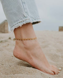 Close-up of a bare foot on sandy beach wearing a gold chain anklet and frayed light-wash jeans hem, beachy summer jewelry shot