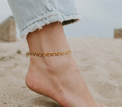 Close-up of a bare foot on sandy beach wearing a gold chain anklet and frayed light-wash jeans hem, beachy summer jewelry shot