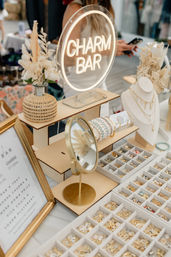 Lighted 'CHARM BAR' sign above a vendor table displaying gold charms, alphabet charms, pastel beaded bracelets and necklace bust at a craft market.