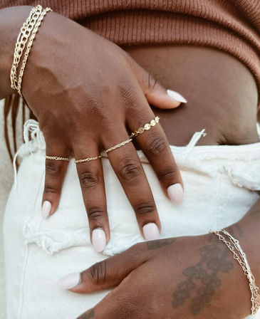 Close-up of hands resting on distressed white denim shorts, showcasing layered gold chain bracelets and rings, pale pink nails and a ribbed brown crop-top — summer street-style accessory detail.