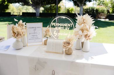 Sunny outdoor vendor table for jewelry with a clear round acrylic sign for permanent jewelry, neutral dried-flower arrangements in textured vases, and a small bracelet display on a white tablecloth.