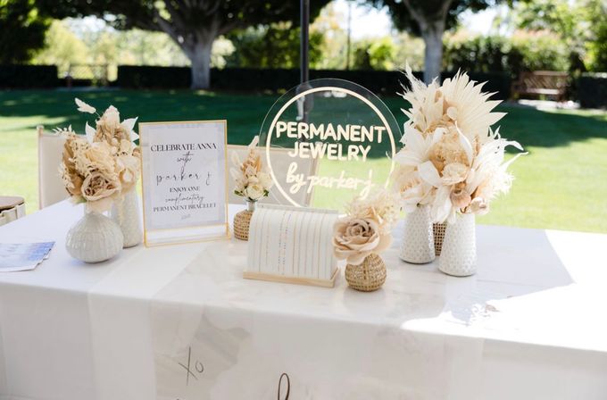 Sunny outdoor vendor table for jewelry with a clear round acrylic sign for permanent jewelry, neutral dried-flower arrangements in textured vases, and a small bracelet display on a white tablecloth.