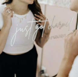 Person adjusting layered gold necklaces in a boutique dressing-room mirror with a playful “just got charmed” script decal, wearing a white crop top and black pants.