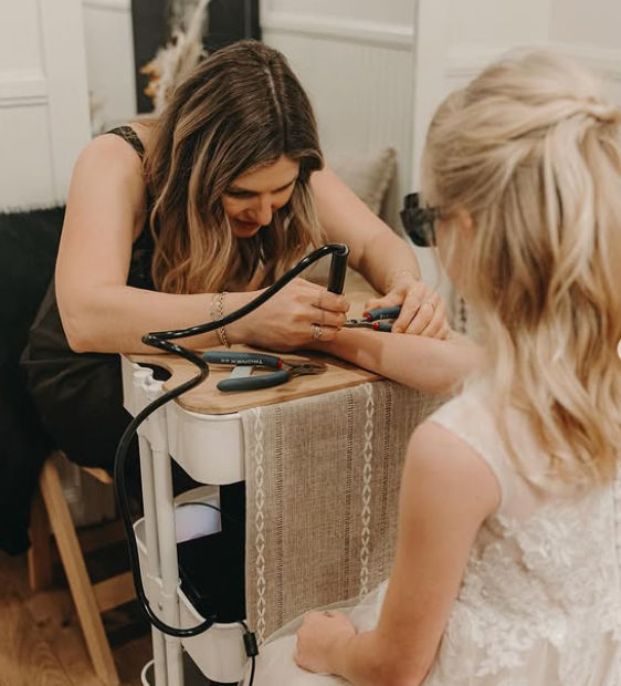 Manicurist painting a young girl's nails on a rolling salon cart during bridal prep in a cozy indoor setting