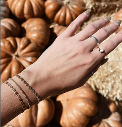 Close-up of a hand with stacked gold bracelets, rings and glittery nails held over orange pumpkins and straw — autumn pumpkin patch accessory shot.