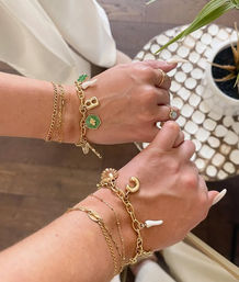Close-up of two wrists wearing layered gold charm bracelets and delicate rings, featuring seashell, crescent moon and green hexagon charms over a wooden floor next to a white mosaic side table with a potted plant.