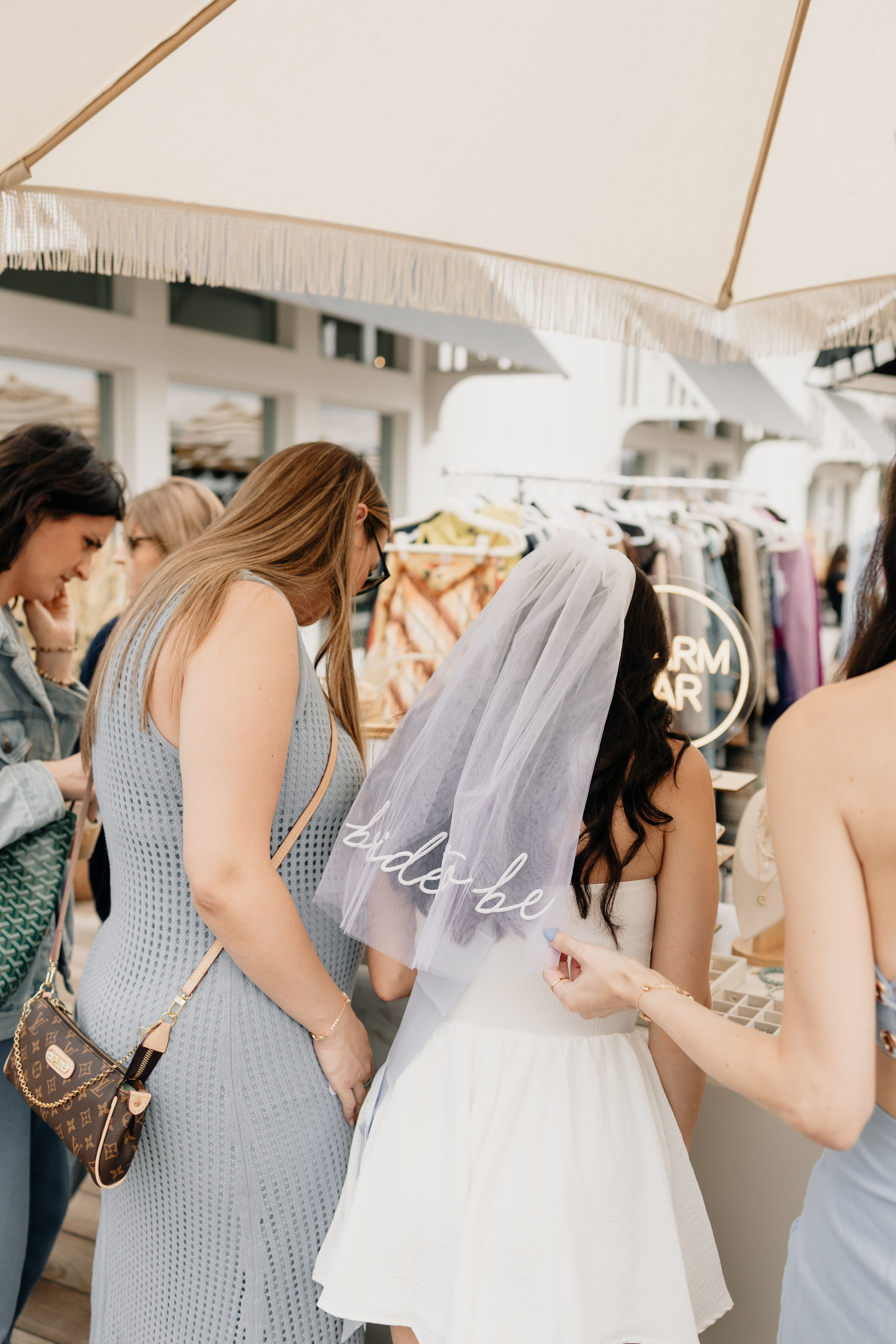 Bride-to-be in a white dress wearing a veil, browsing market stalls with friends under a fringed umbrella — outdoor bridal shower shopping scene with clothing and jewelry racks.