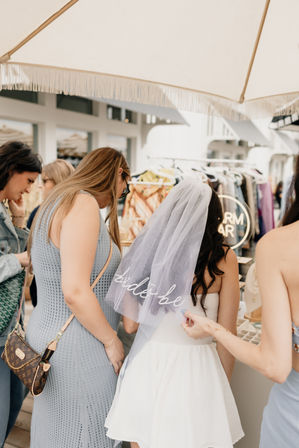 Bride-to-be in a white dress wearing a veil, browsing market stalls with friends under a fringed umbrella — outdoor bridal shower shopping scene with clothing and jewelry racks.