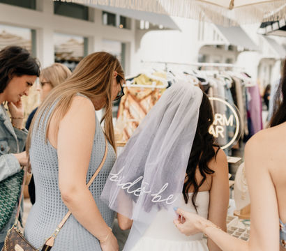 Bride-to-be in a white dress wearing a veil, browsing market stalls with friends under a fringed umbrella — outdoor bridal shower shopping scene with clothing and jewelry racks.