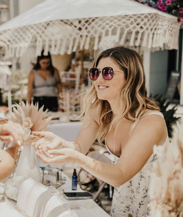 Smiling woman vendor in sunglasses hands over jewelry at a sunny outdoor market stall under a fringed sun umbrella with boho displays and dried floral decor.