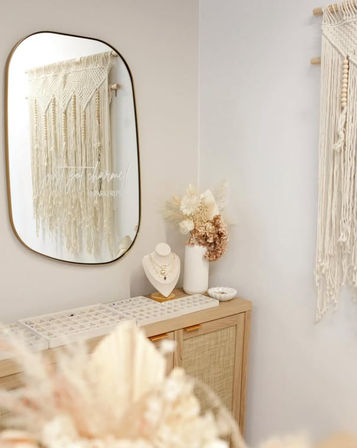 Cozy chic boho corner with macramé wall hangings, oval mirror, rattan cabinet topped with a jewelry display tray, necklace bust, and dried floral arrangement.