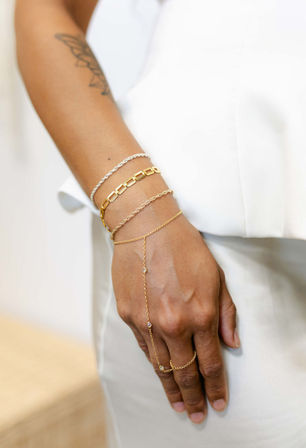 Close-up of a hand and wrist wearing stacked gold and silver bracelets and a delicate gold hand-chain ring styled against a white outfit — dainty minimalist women's jewelry.