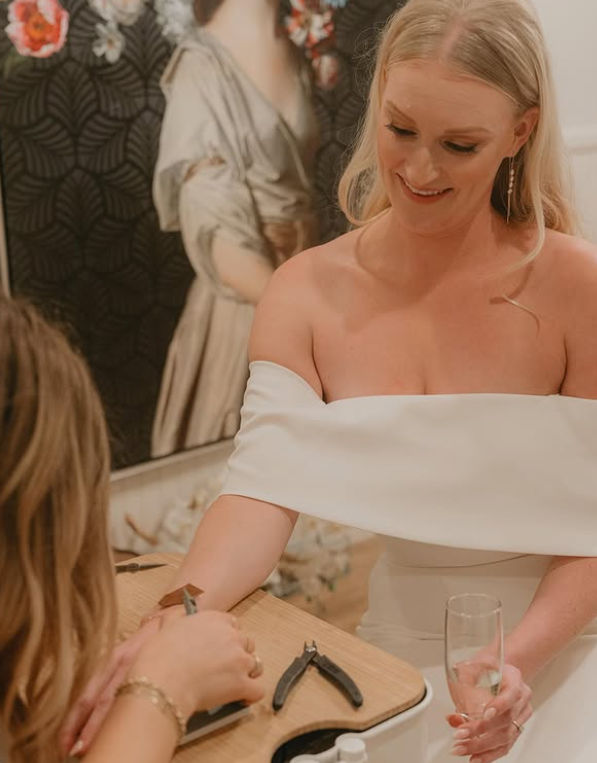 Smiling bride in an off-the-shoulder white dress holding a champagne flute while receiving a manicure at a salon table with nail tools, wedding-day pampering.